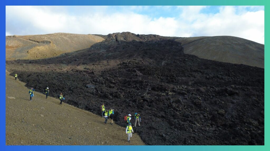 A group of people in high-vis jackets are spread out along a lava deposit in a wide landscape.